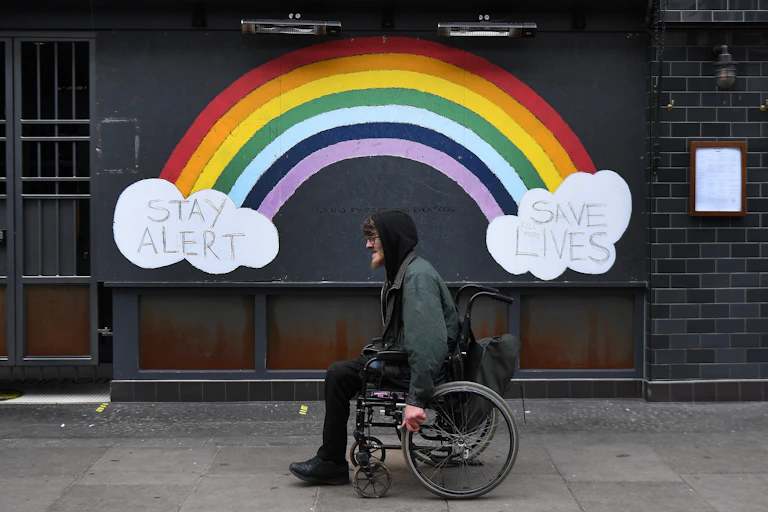 A man in a wheelchair passes a street mural of a rainbow and the slogan "stay alert save lives".