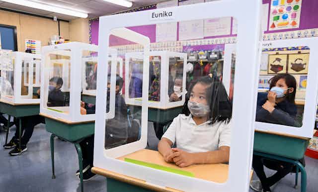 Children wearing masks behind screens in a classroom.