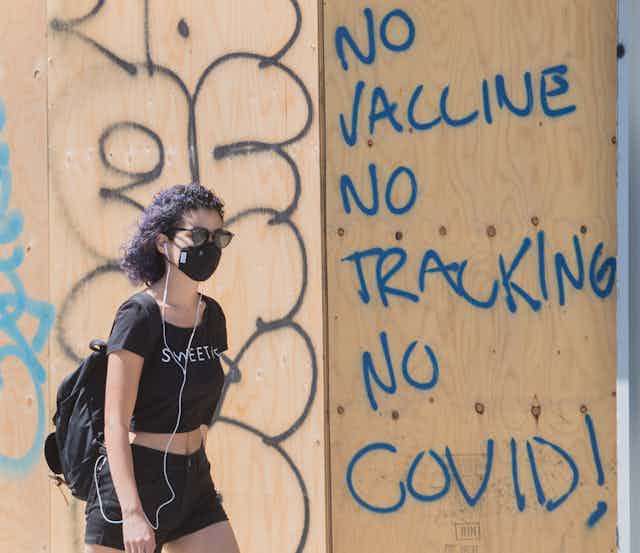 A woman wearing a face mask walks by a board-up window with 'no vaccine no tracking no COVID!' written on it in blue letters.