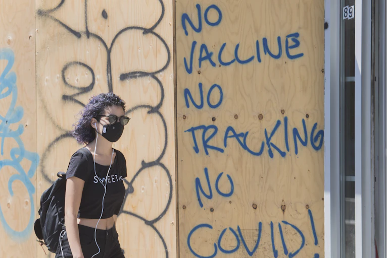 A woman wearing a face mask walks by a board-up window with 'no vaccine no tracking no COVID!' written on it in blue letters.