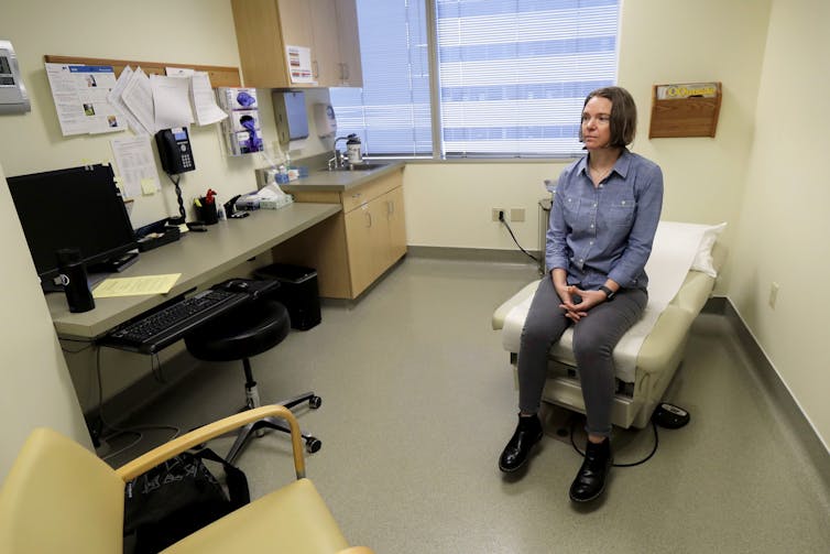 A woman in jeans and a denim shirt sits on the end of an exam table in a medical exam room.