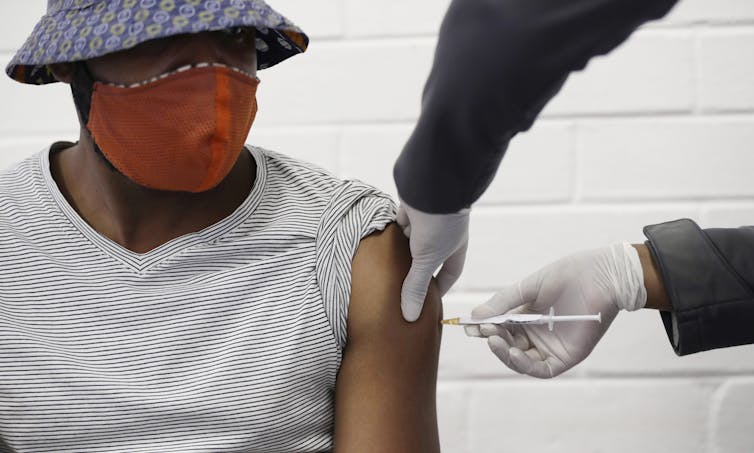 A man wearing a hat and a face mask gets an inject from a health-care worker out of frame.