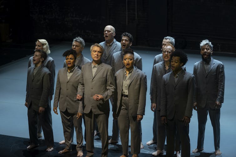 A group of people in grey suits sing out towards the audience.