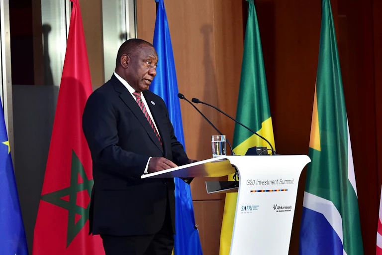 A man wearing a dark suit and striped maroon and black tie speaks at a podium, flanked by flags of various countries.