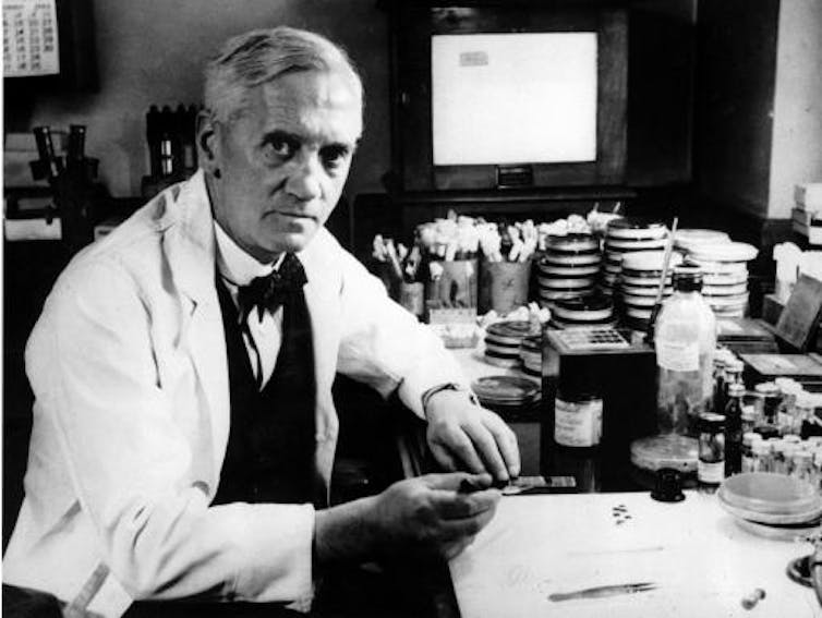 Alexander Fleming seated, wearing a white lab coat.