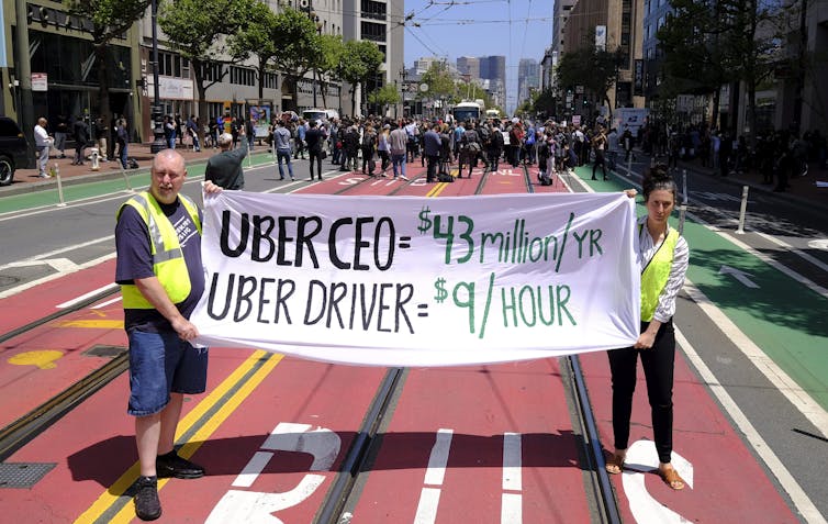 Two people hold up a sign decrying low wages for Uber drivers on a city street.