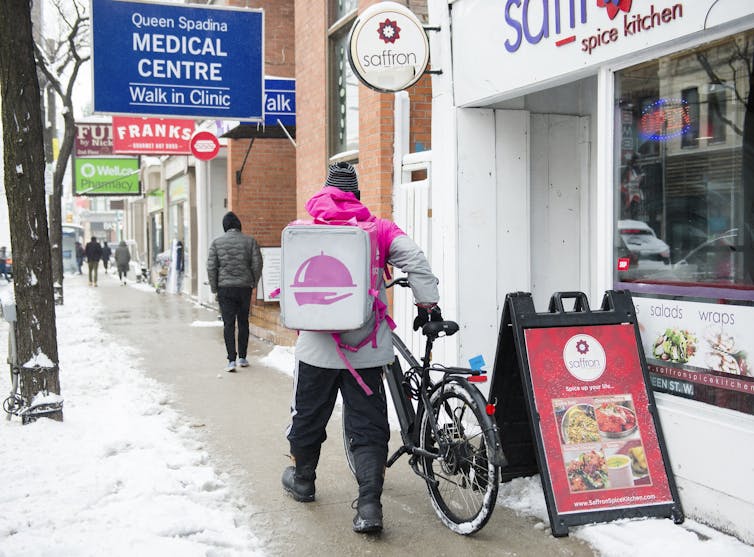 A Foodora courier with a food order on his back pushes his bicycle along a snowy street.