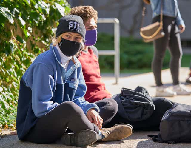 Two casually dressed young people in face masks sit on the ground with their backpacks beside them.