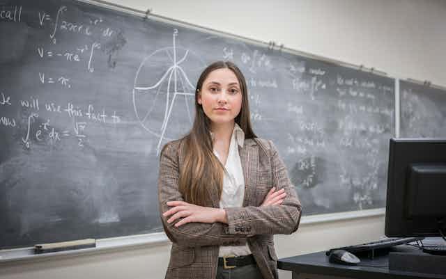 Young college instructor stands with arms folded in fron of math equations on a chalkboard.