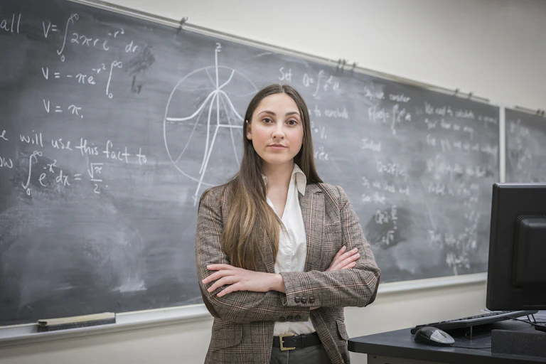 Young college instructor stands with arms folded in fron of math equations on a chalkboard.