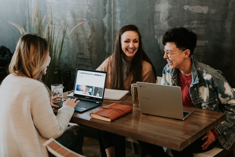 A man and two women sit at a table with their laptops chatting and laughing.