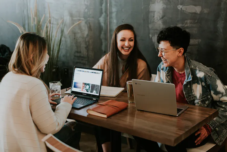 A man and two women sit at a table with their laptops chatting and laughing.