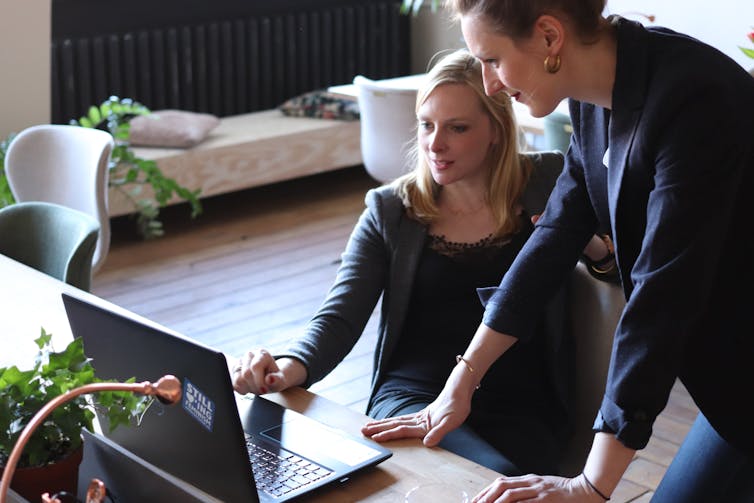 A woman sits at a laptop as another woman stands beside her looking at her screen.