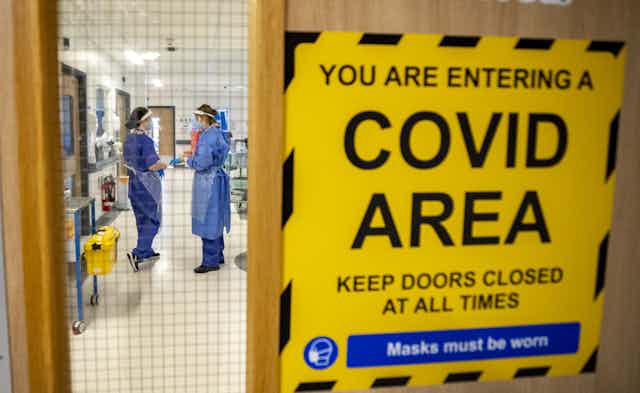 Two healthcare workers in full PPE seen through a door bearing a sign saying "YOU ARE ENTERING A COVID AREA"