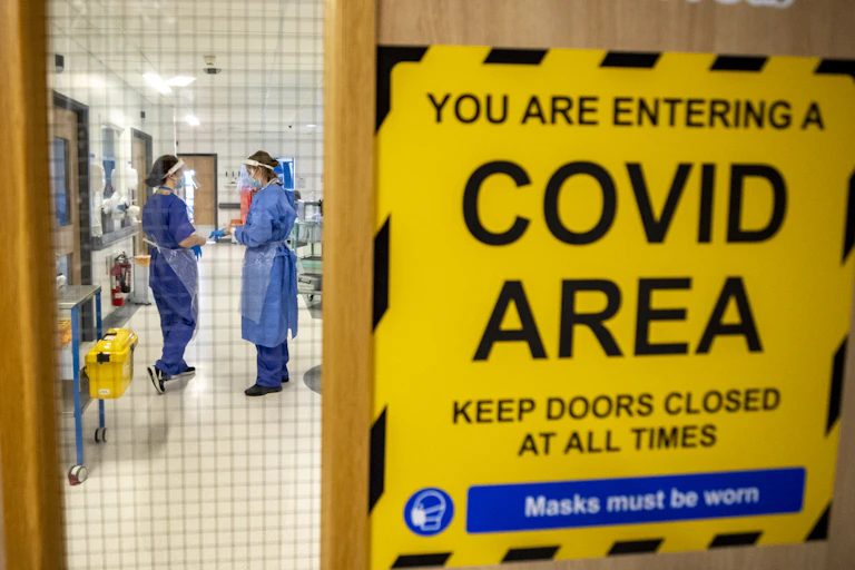 Two healthcare workers in full PPE seen through a door bearing a sign saying "YOU ARE ENTERING A COVID AREA"