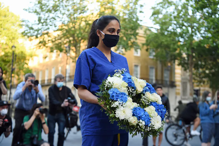 A woman wearing hospital scrubs and a mask carries a wreath of flowers.