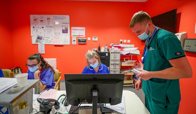 Three healthcare workers in scrubs gather around a desk in a hospital.