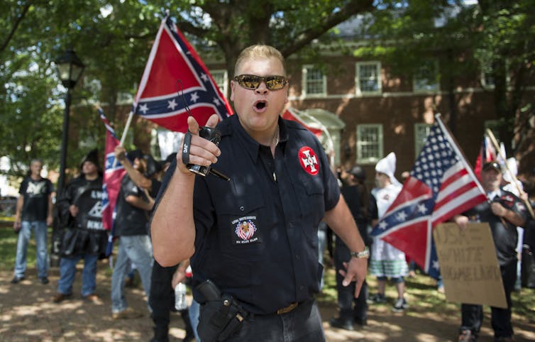A man wearing white supremacist symbols and a holstered pistol points while several people stand behind him waving Confederate flags