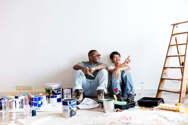 a couple sit on the floor surrounded by paint cans next to a ladder