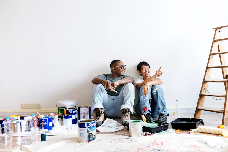 a couple sit on the floor surrounded by paint cans next to a ladder