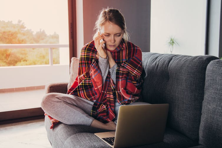 A woman sits on a couch, wrapped in a blanket, speaking on the phone. She has a laptop in front of her.