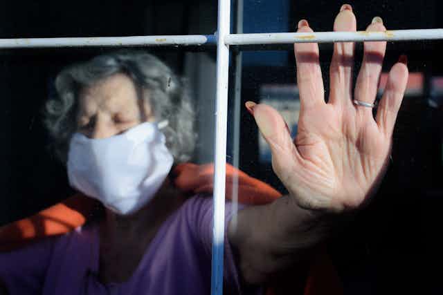 Una señora mayor con mascarilla, tras una ventana, apoya la palma de la mano en el cristal.