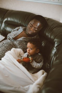 A man is asleep on the sofa with his small child who is awake watching something.