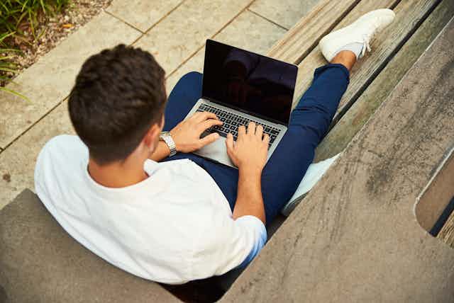 A man sits on the floor on his laptop in a white jumper and blue trousers