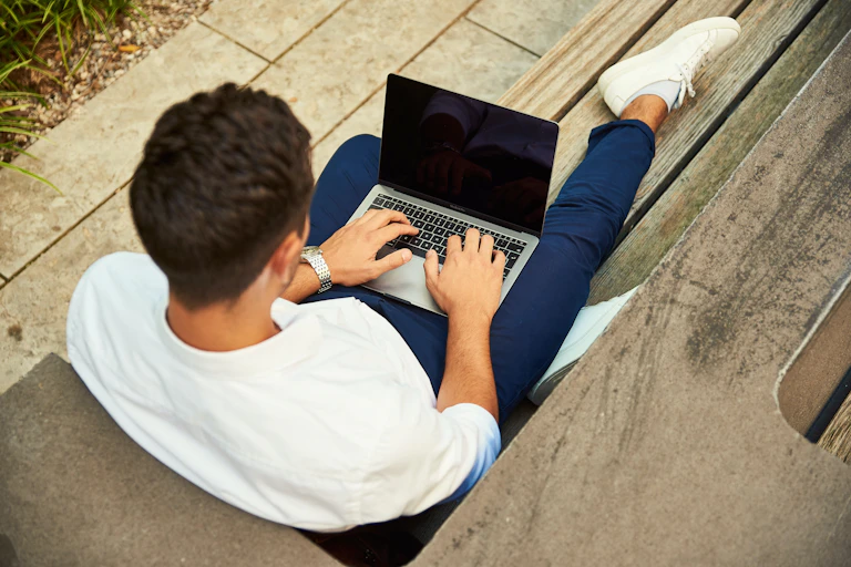 A man sits on the floor on his laptop in a white jumper and blue trousers