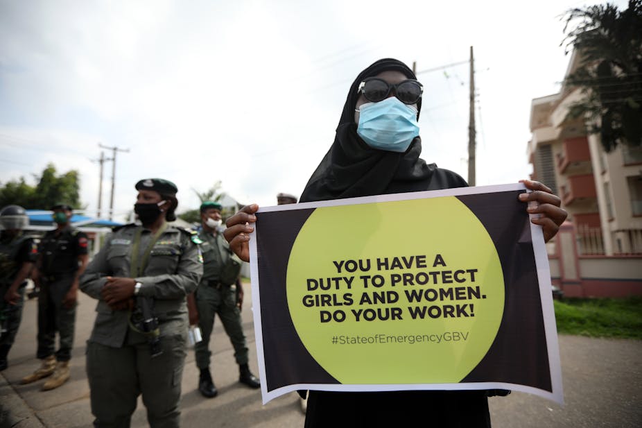 Woman wearing a mask holds a placard, while police officers stand behind her.