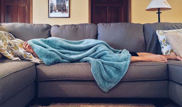 A person lying a sofa, almost entirely covered by a fluffy blue blanket