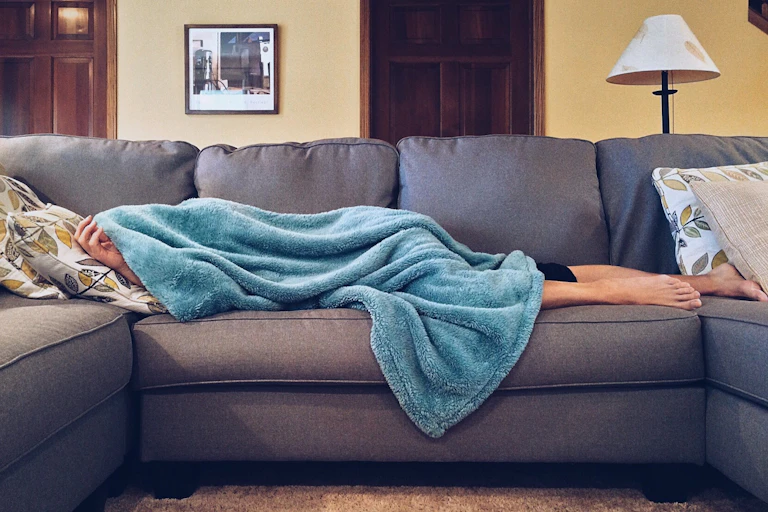 A person lying a sofa, almost entirely covered by a fluffy blue blanket
