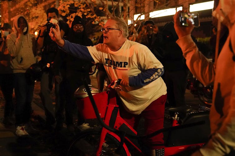A man on a bicycle in a Trump T-shirt sprays something at someone standing nearby
