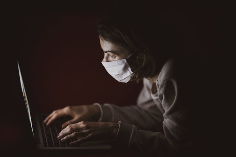 Person in mask looks at laptop in darkened room