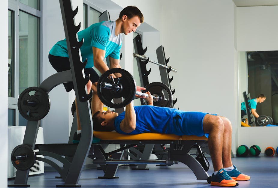 A male gym trainer helping a young man perform a bench press.