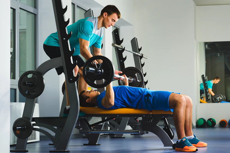 A male gym trainer helping a young man perform a bench press.