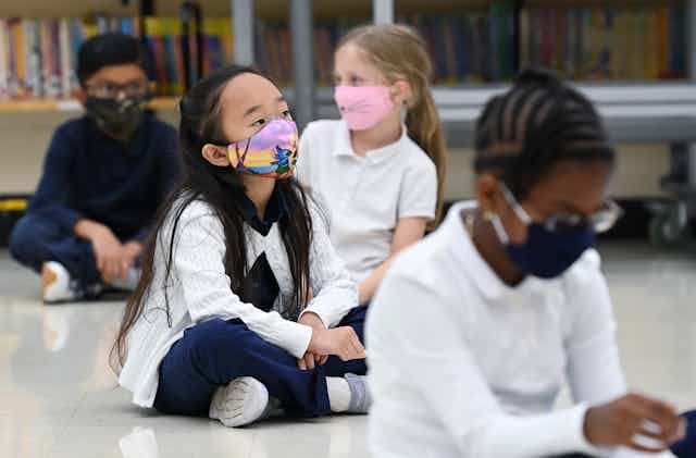 Children sit cross-legged on the floor at school wearing masks.