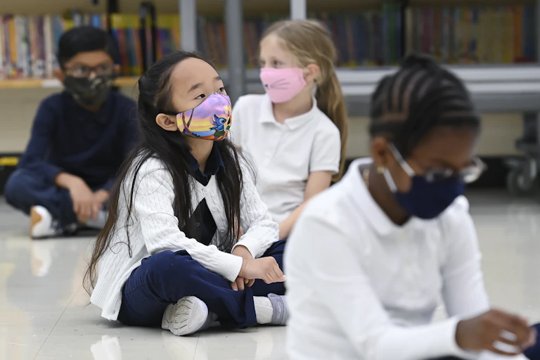 Children sit cross-legged on the floor at school wearing masks.