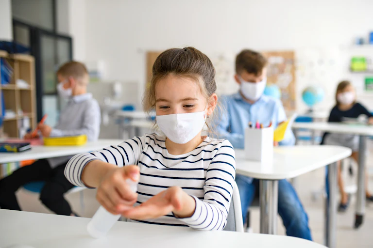 Cuatro niños con mascarilla en un aula. En primer plano una niña se echa gel hidroalcohólico en las manos.