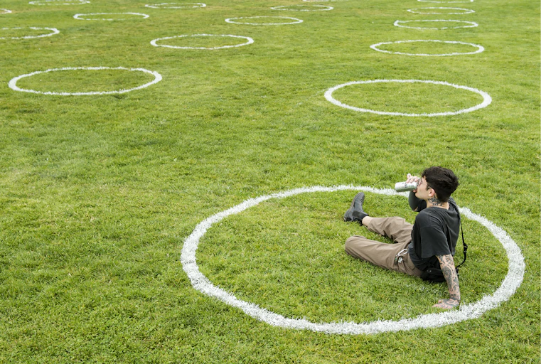 A man sits on the grass in a white circle, with dozens of empty white circles on the expanse of grass in the background