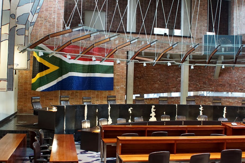 Interior of courtroom showing seating for judges, benches for public, and South African flag on the wall