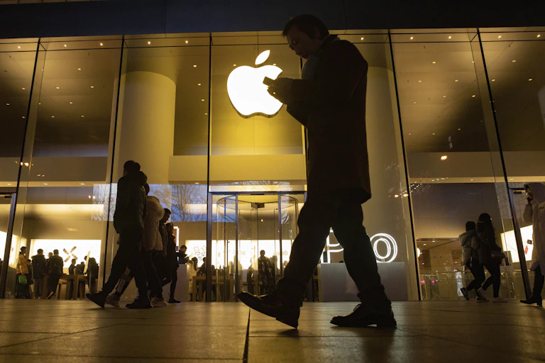A man stares down at his phone while walking past an Apple store.