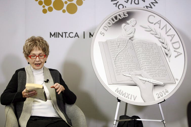 The author Alice Munro reads from her book while sitting next to a large replica of the $5 coin celebrating her achievements.