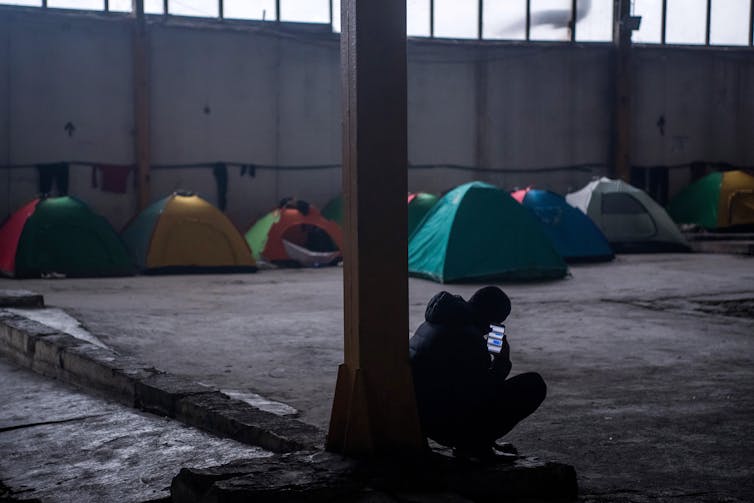 Silhouette of man crouching with mobile phone in hand with tents in the background