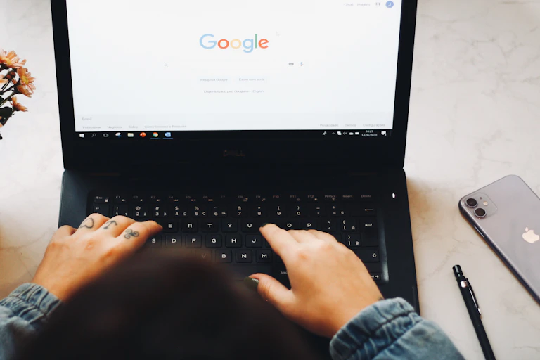 Two tattooed hands type on a laptop with Google on the screen with an apple phone next to them and a bunch of flowers