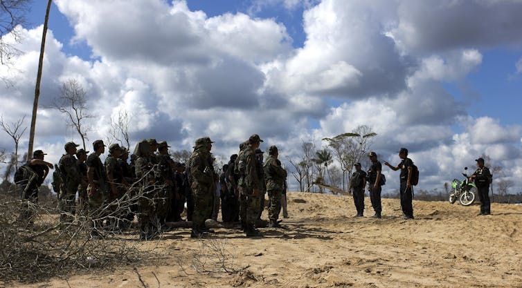 Police officers in fatigues stand on pitted, sandy ground