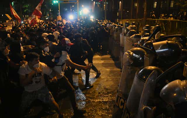 Police in riot gear with shields stand off against unarmed protesters holding signs and flags at night