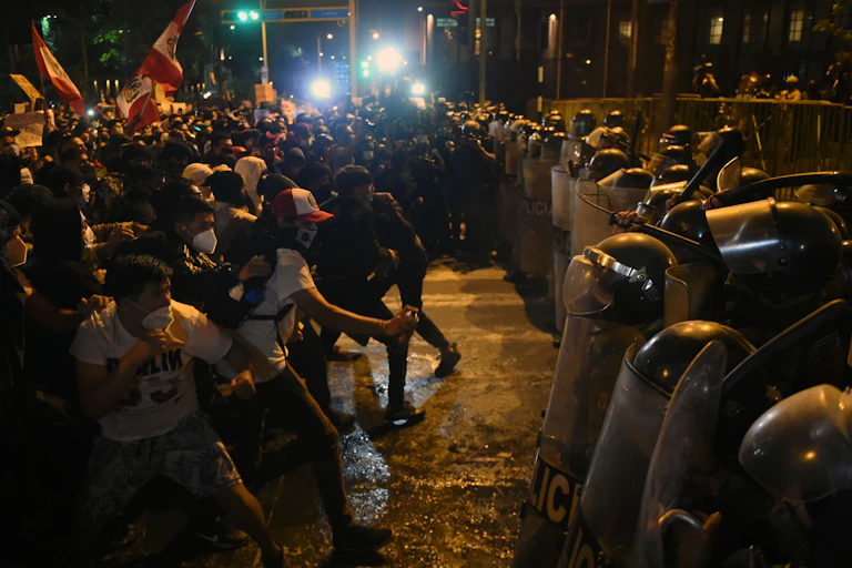 Police in riot gear with shields stand off against unarmed protesters holding signs and flags at night