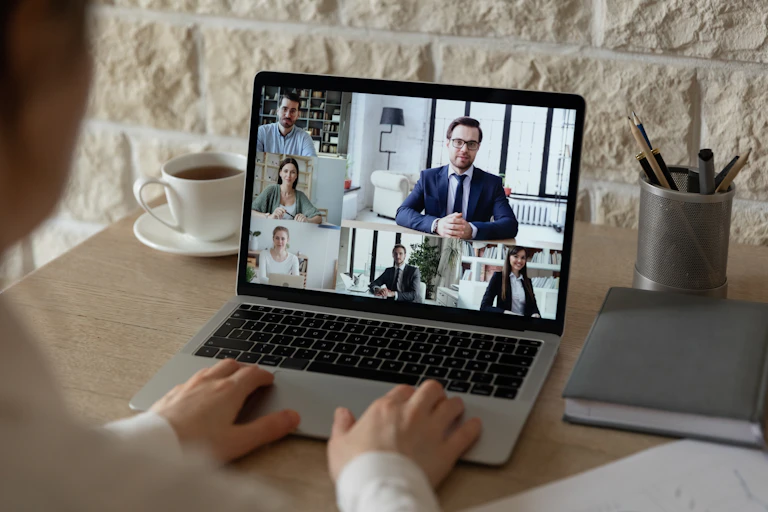 Hands on a laptop showing six people in business attire in a videoconference.