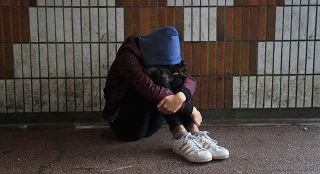 Teen wearing maroon jacket, blue hoodie and sneakers sits with her head between her knees